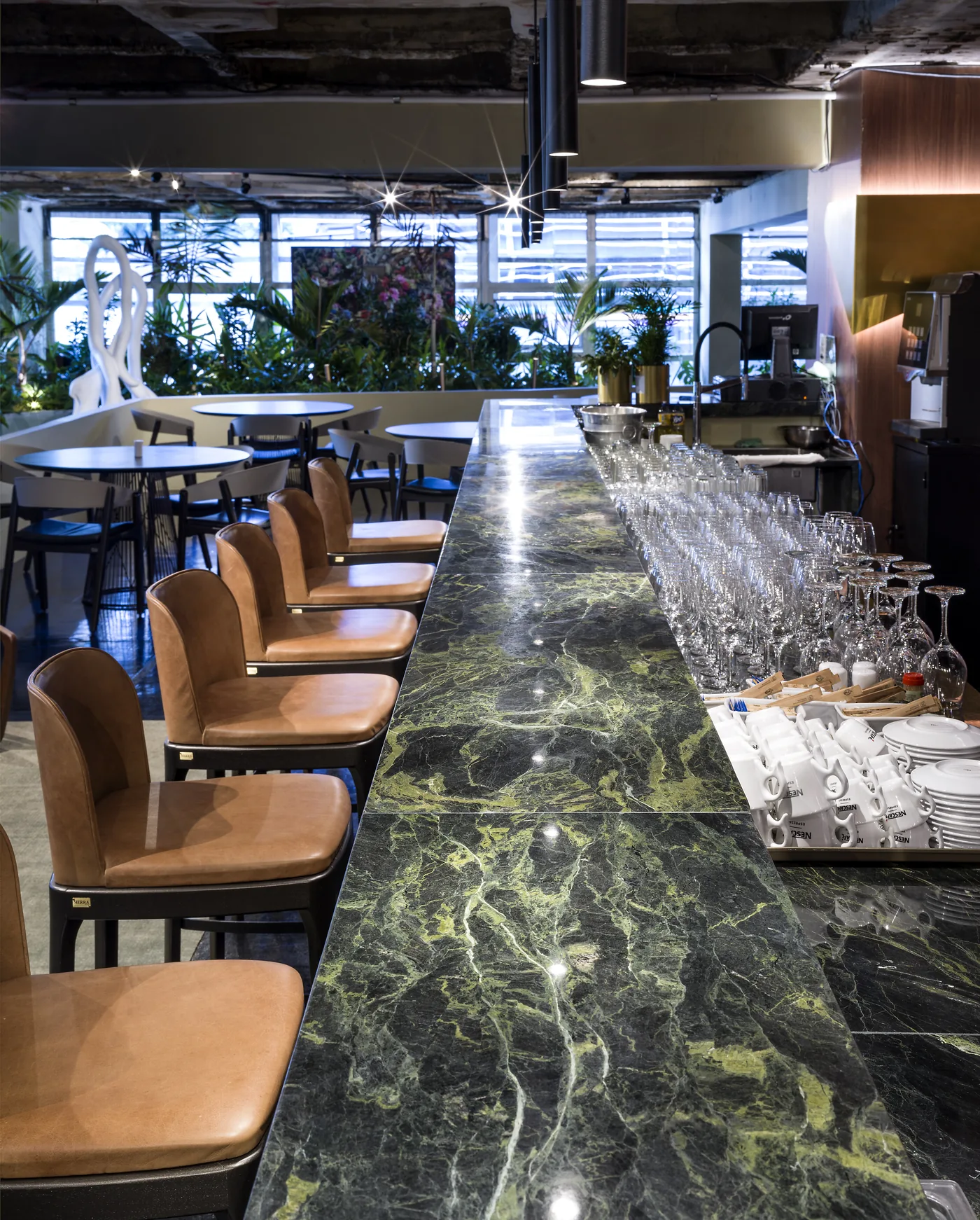 Bancada do bar com copos organizados, mármore verde e salão ao fundo