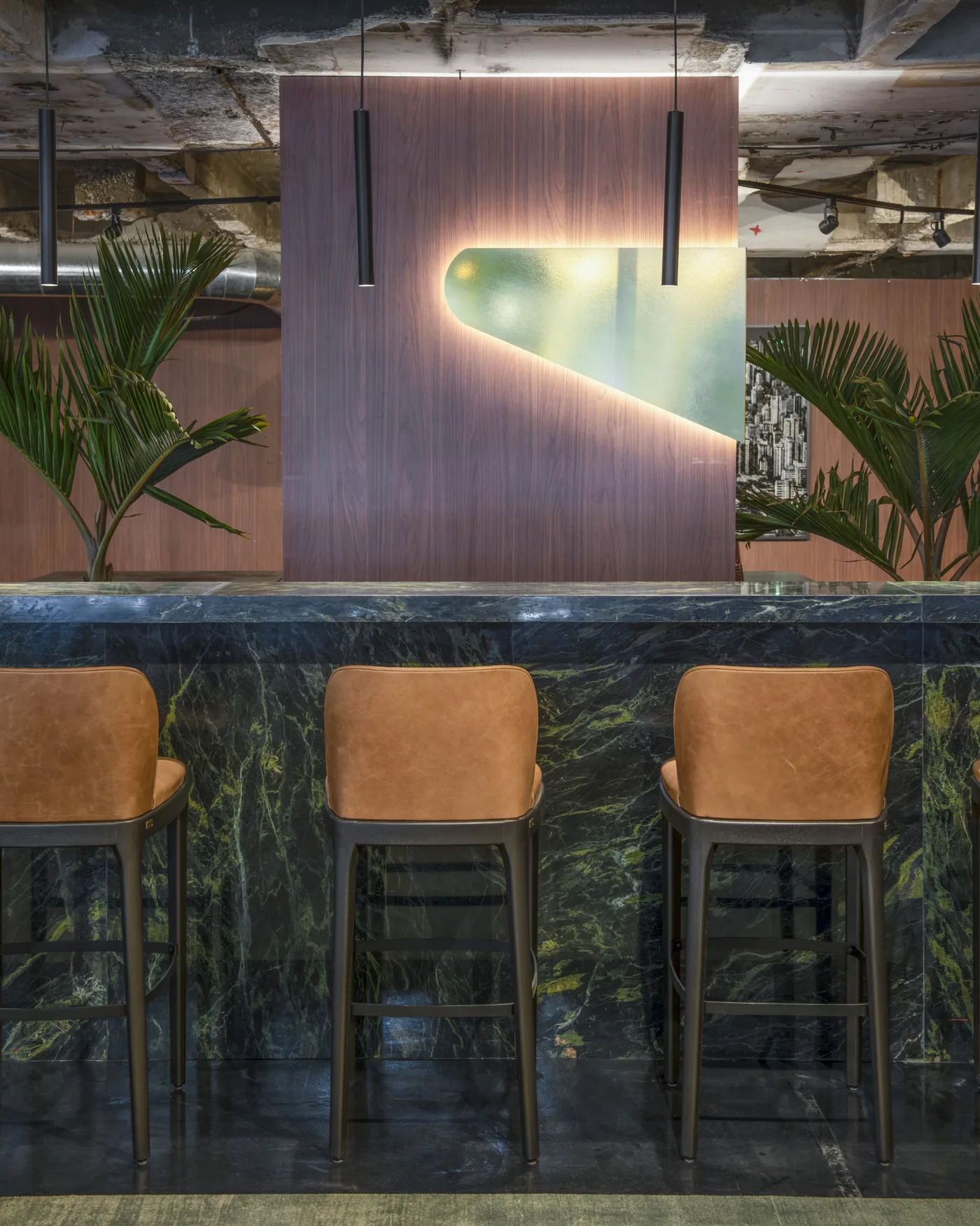 Detalhe do bar com painel verde iluminado, mármore escuro e banquetas de couro