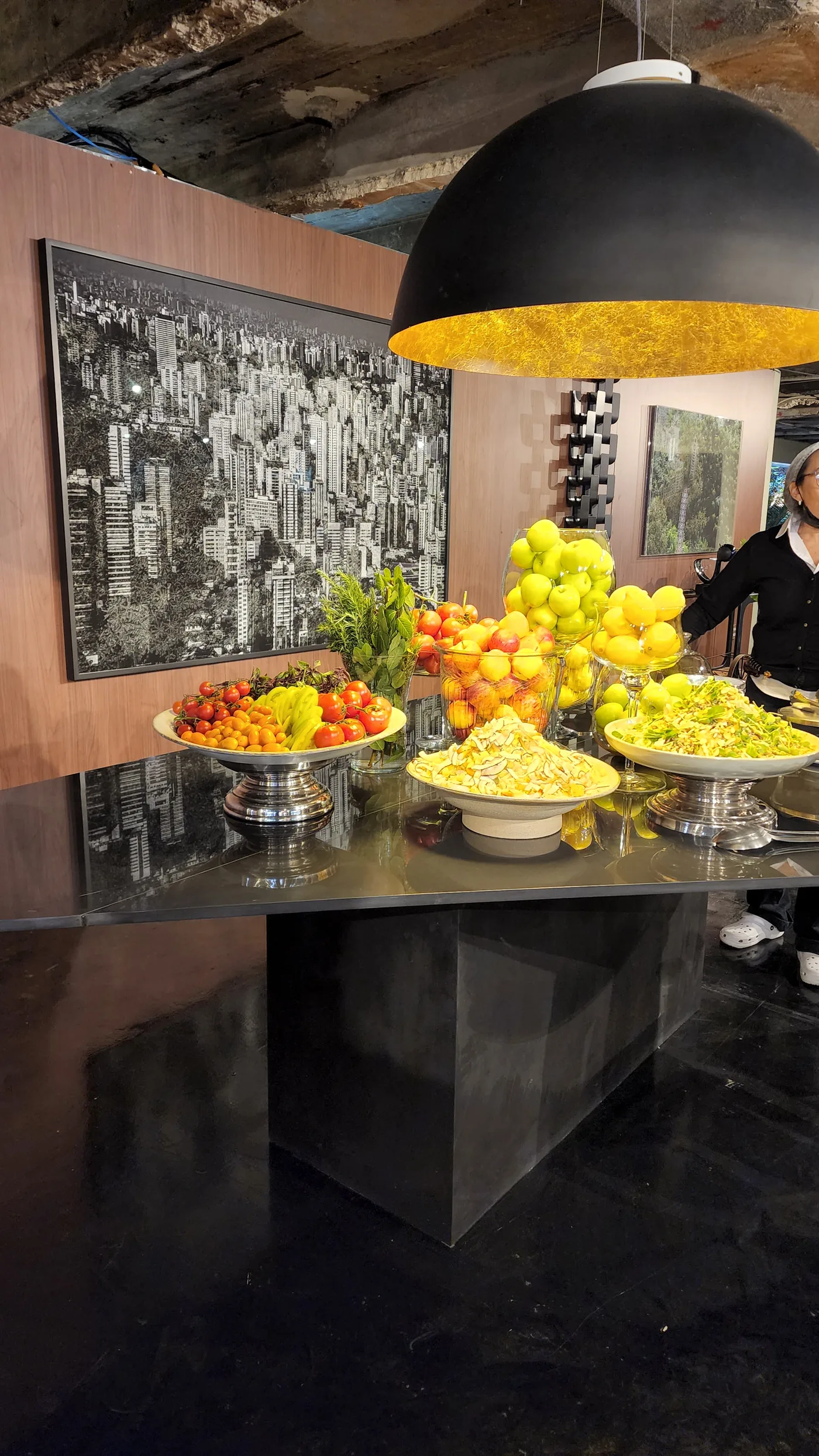 Mesa de apoio com frutas, pendente preto e dourado e fotografia urbana ao fundo