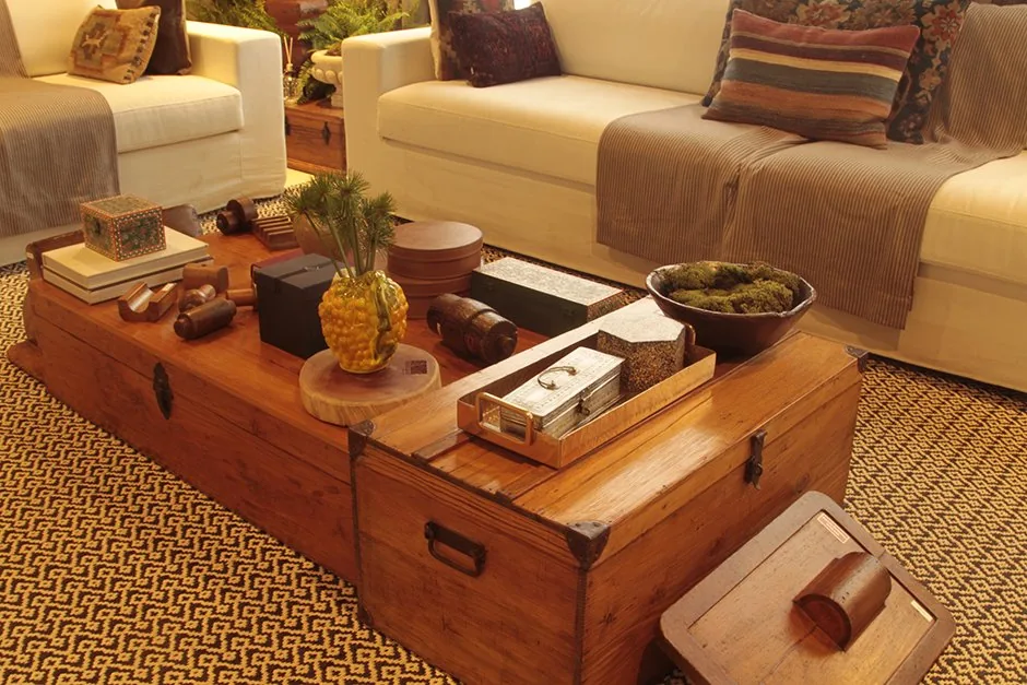 Detalhe da mesa de centro em madeira com livros, objetos decorativos e sofás brancos ao fundo
