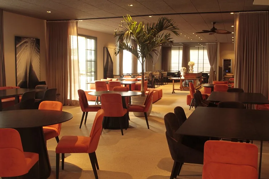 Vista geral do CASACOR BADEBEC Living do Restaurante com mesas, poltronas laranja e palmeira central