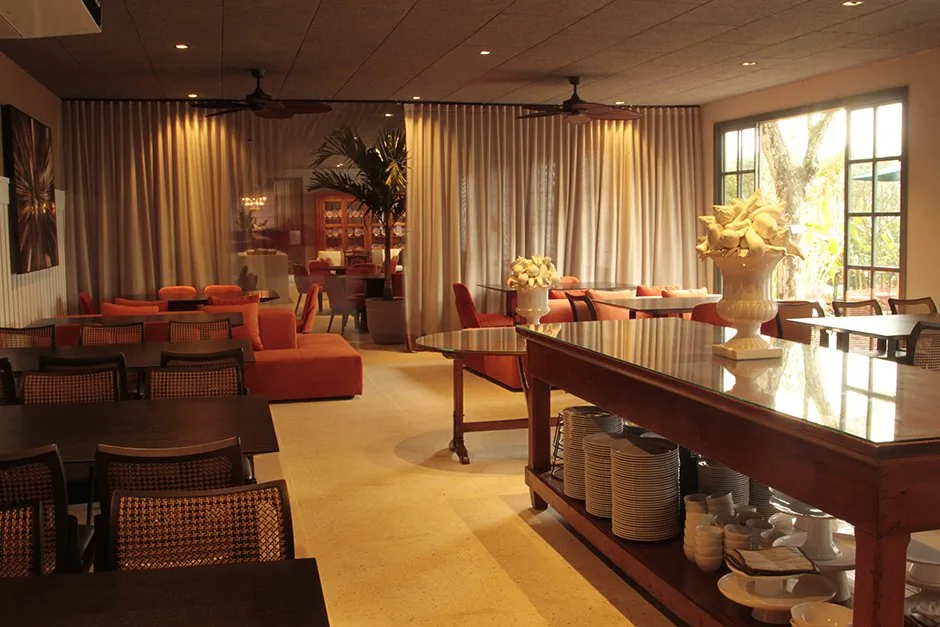 Vista do CASACOR BADEBEC Living do Restaurante com mesas, poltronas laranja, buffet e cortinas
