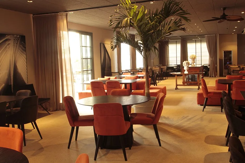 Salão do restaurante com poltronas laranja, palmeira central e mesas distribuídas