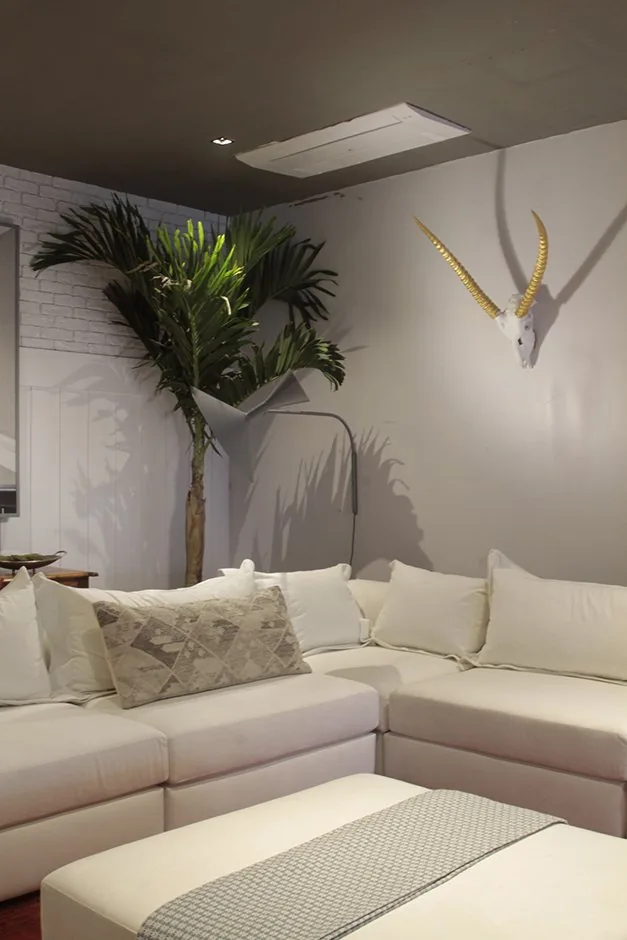 Sofá branco de canto com almofadas claras, palmeira e parede decorada com cabeça de antílope