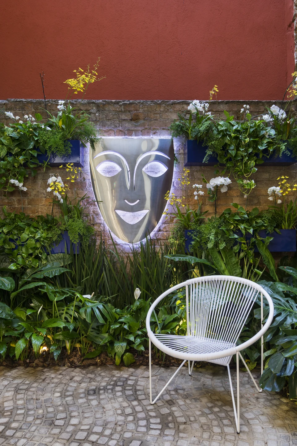 Jardim interno da Kryolan Flagship com máscara metálica iluminada e paisagismo tropical.